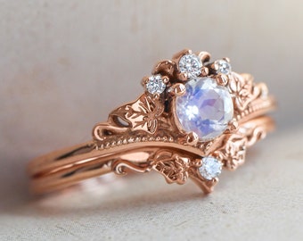 May include: Rose gold engagement ring set with a round, iridescent moonstone center stone and small, clear accent stones. The ring features a floral and leaf design, with intricate details. The band is also rose gold.