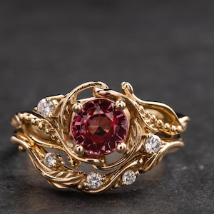 May include: A gold ring with a red gemstone surrounded by smaller diamonds. The ring features a delicate, intricate design with a floral motif.
