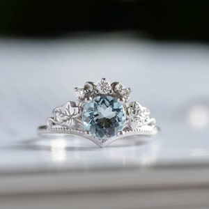 May include: A silver ring with a light blue aquamarine gemstone surrounded by smaller diamonds. The ring has a delicate floral design.