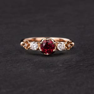 Peut inclure: Une bague en or rose avec une pierre précieuse en rubis rouge et deux petits diamants blancs sertis dans un motif floral.