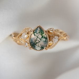 Può includere: Un anello d'oro con una pietra preziosa di agata verde muschio a forma di pera, circondata da diamanti più piccoli. L'anello presenta un design delicato e fogliare.