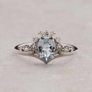 Può includere: Un anello d'argento con una pietra preziosa di acquamarina a forma di pera, decorato con un delicato motivo floreale e piccoli diamanti.