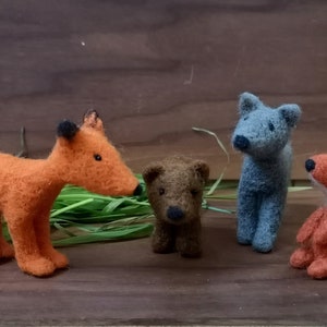 Pode incluir: Quatro figuras de animais em lã feltrada, incluindo uma raposa laranja grande, um urso castanho pequeno, um lobo cinzento de pé e uma raposa laranja pequena com a ponta da cauda branca. As figuras estão alinhadas com fios de erva verde.