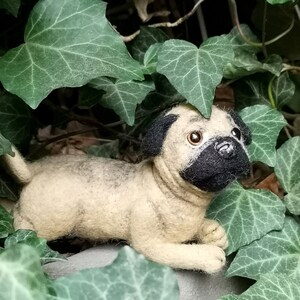 May include: A needle-felted pug figurine featuring a light tan body, a black muzzle, and black ears. The small dog sculpture has dark brown eyes and a shiny black nose, crafted with a textured wool finish to resemble a realistic pug dog.