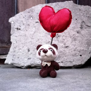 May include: A handcrafted panda bear figure holding a red heart-shaped balloon. The panda has a white face, brown ears and body, and a small gold bow tie. The balloon is a deep red colour.