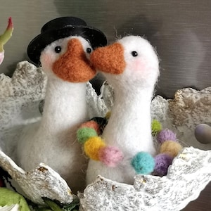 Può includere: Due statuette di oche bianche con becchi arancioni e occhi neri. Un'oca indossa un cappello nero, l'altra ha una collana di pompon colorati. Sono adagiate in un nido decorativo e strutturato.