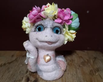 Könnte beinhalten: Ein rosa Filzwurm mit einer Blumenkrone, blauen Augen und einem rosa Herz-Anhänger um den Hals. Der Wurm sitzt auf einer Holzoberfläche.