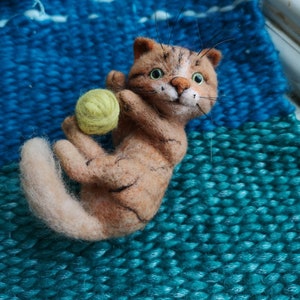 May include: A needle-felted tan tabby cat figurine with dark brown stripes, green eyes, and long black whiskers. The cat is holding a small, round, light green ball of yarn between its front paws, with a curved tail and a white muzzle.