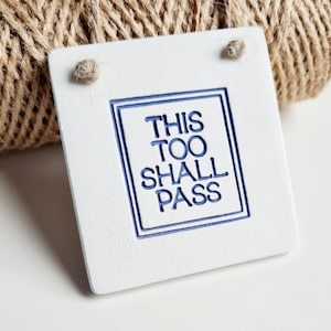 Puede incluir: Etiqueta de cuero blanco con borde azul y el texto "This Too Shall Pass" en letras azules.
