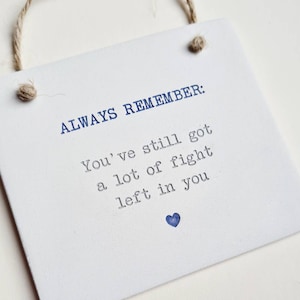 Może przedstawiać: Biała tabliczka z niebieskim tekstem, który brzmi: "ALWAYS REMEMBER: You've still got a lot of fight left in you." Małe niebieskie serce znajduje się na dole tekstu.
