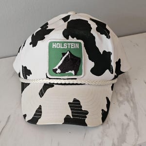 May include: White and black Holstein cow print baseball cap with a green patch that reads "Holstein" and features a cow graphic. The cap has a white rope detail and a curved brim.