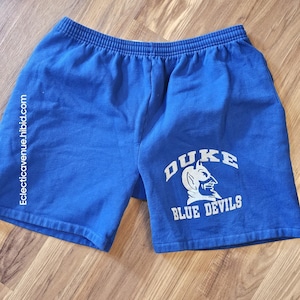 Könnte beinhalten: Königsblaue Sporthose mit elastischem Bund. Die Shorts zeigen den Text "DUKE BLUE DEVILS" in Weiß, zusammen mit einer Teufel-Maskottchen-Grafik. Die Website "Eclecticavenue.hibid.com" ist vertikal auf dem linken Bein aufgedruckt.