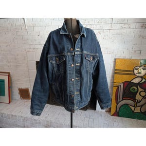 Könnte beinhalten: Dunkelblaue Jeansjacke mit klassischem Design. Die Jacke hat eine Knopfleiste vorne, zwei Brusttaschen und zwei Seitentaschen. Die Jacke wird auf einer Schaufensterpuppe vor einer weißen Backsteinmauer präsentiert.