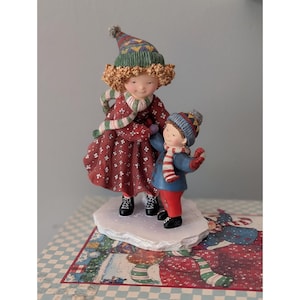 May include: A figurine of two figures standing on a white base. The taller figure wears a red dress with white floral patterns, a green and white striped scarf, and a multi-colored hat. The smaller figure wears a blue coat, red pants, and a blue hat.