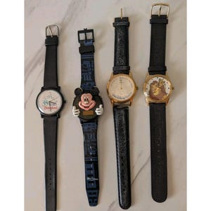 May include: Four vintage Disney watches with black leather bands. One watch features Mickey Mouse, another has a 35th Anniversary Disneyland design, and two others have gold-toned faces with Disney characters. The watches are displayed on a light-colored surface.