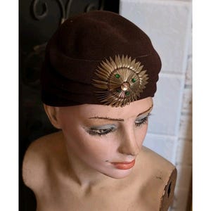 May include: A brown vintage hat with a decorative gold and green embellishment. The hat has a rounded shape with a pleated design. The gold embellishment is circular with green accents.