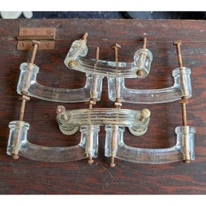May include: Four clear glass drawer pulls with a curved, ribbed design. Each handle features two long, threaded metal bolts that show significant rust and oxidation. The pulls are arranged in a rectangular formation, showcasing the transparent glass.
