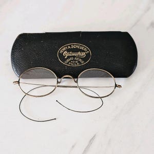 Könnte beinhalten: Vintage runde Brille mit goldfarbenem Rahmen und schwarzem Etui. Auf dem Etui steht "Henry A. Scheueri Optometrist, Girard Avenue, Phila."