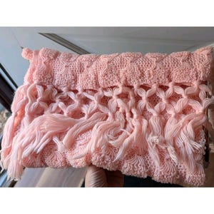May include: A light pink knitted scarf with a textured pattern and fringe detailing. The scarf is made of soft yarn and has a delicate, feminine appearance. The fringe adds a playful touch to the design.