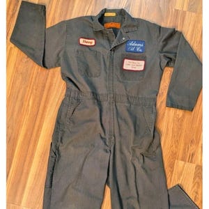 May include: A charcoal grey long-sleeve coverall featuring a collar, button-up front, and two chest pockets. Patches include a red name tag reading Harry, a blue patch reading Adams Oil Co., and a white rectangular patch with red text.