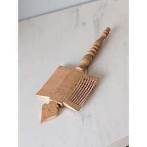 May include: A light brown wooden comb with a long, turned handle and a decorative, carved base. The comb features fine teeth and a detailed floral design on the base. The comb is likely vintage or antique.