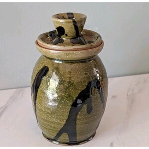 Pode incluir: Um pote de cerâmica com tampa, apresentando um esmalte verde-oliva salpicado e padrões abstratos de gotas em castanho escuro. O pote tem um corpo arredondado e uma tampa a condizer com um puxador central, com um acabamento liso e brilhante.