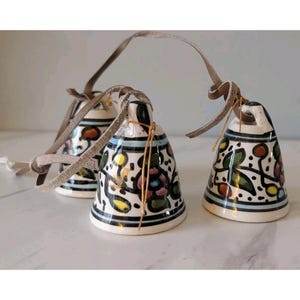 May include: Three ceramic bells with a floral design in various colors, including orange, green, and yellow, are suspended by leather straps. The bells have a black and white striped pattern at the top and bottom.