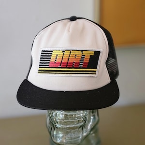 Puede incluir: Gorra de camionero negra y blanca con un panel frontal blanco. El panel frontal presenta un parche rectangular con la palabra "DIRT" en un diseño retro multicolor. La visera es negra.