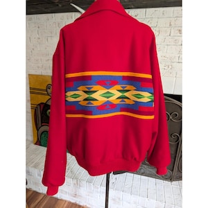 May include: A bright red jacket featuring a wide horizontal band across the back with a geometric pattern in blue, yellow, green, and orange. The jacket has a collared neckline, long sleeves with ribbed cuffs, and a ribbed hem at the bottom edge.