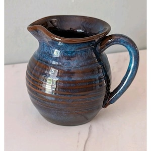 May include: A ceramic pitcher featuring a rounded body with horizontal ribbed textures. The glaze displays a blend of deep brown and vibrant blue tones, with a glossy finish. It includes a curved handle and a wide, shaped pouring spout.