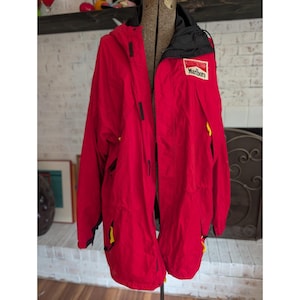 May include: A bright red hooded jacket featuring a black collar and interior lining. The jacket has a white rectangular patch on the chest with the red and black text Marlboro. It includes black vertical closures and small yellow accents on the pockets.