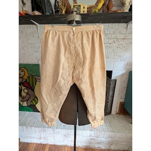 May include: A pair of light beige, loose-fitting trousers featuring a high-waisted design with a single centered button closure at the waistband. The pants have a relaxed, wide-leg silhouette that tapers slightly toward the gathered, elasticized cuffs.