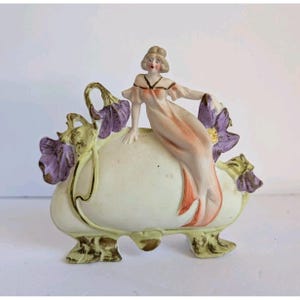 Op de afbeelding: Een porseleinen beeldje met een zittend figuur in een perzikkleurige japon op een crèmekleurige ovale basis. De basis is versierd met gebeeldhouwde paarse bloemen en groene bladeren, ondersteund door vier sierlijke, goudkleurige voetjes.
