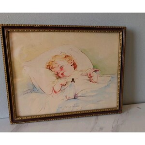 May include: Framed vintage print of a sleeping baby with blonde curls, resting on a white pillow and blanket. The artwork features soft watercolor hues and the text "PILLOW OF DREAMS". The frame is a dark brown with gold accents.