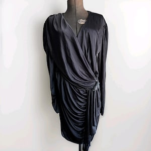 May include: A black, long-sleeved dress with a draped, wrap-style design. The dress features a V-neckline and a gathered, asymmetrical skirt. A decorative element is at the waist. The dress is displayed on a black dress form against a white background.