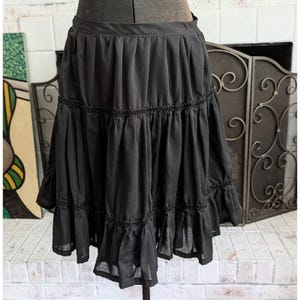 May include: A black knee-length skirt featuring a gathered waistband, two horizontal tiers of fabric, and decorative black trim detailing along the seams. The skirt is constructed with a lightweight, semi-sheer fabric that creates a full, pleated silhouette.