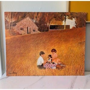 May include: Vintage print depicting a picnic scene with two children and a dog in a field of tall grass. A stone house and trees are in the background. The children are sitting on a red and white checkered blanket with a picnic basket.