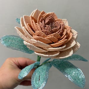 May include: A handcrafted flower sculpture made from tiny glass beads. The central petals transition from a deep terracotta to a soft cream color. The stem and pointed leaves are constructed from vibrant turquoise beads, creating a contrast.