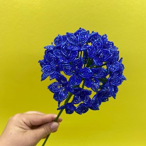 May include: A blue beaded flower with a green stem. The flower is made of small, round beads that are arranged in a flower shape. The stem is made of wire and is wrapped in green tape.