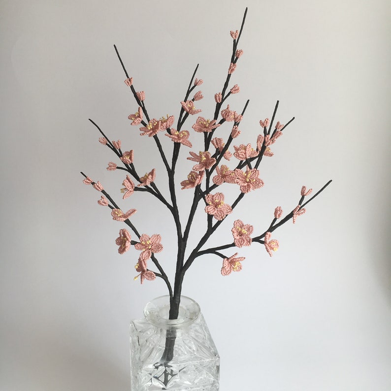 May include: A delicate pink beaded flower arrangement in a clear glass vase. The flowers are arranged in a branch-like shape, creating a whimsical and elegant display.