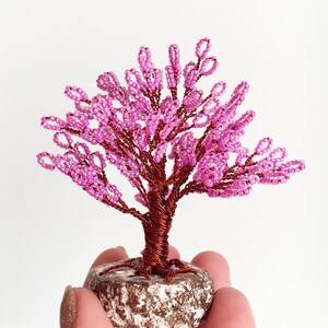May include: A pink beaded wire tree sculpture with a brown wire trunk and a white base.
