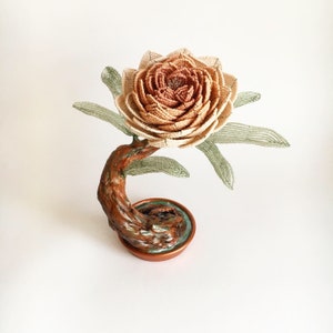 May include: A handcrafted, beaded flower sculpture with a detailed, layered petal design in shades of beige and brown. The flower sits atop a curved, textured, brown stem and a copper-colored base with a touch of green.