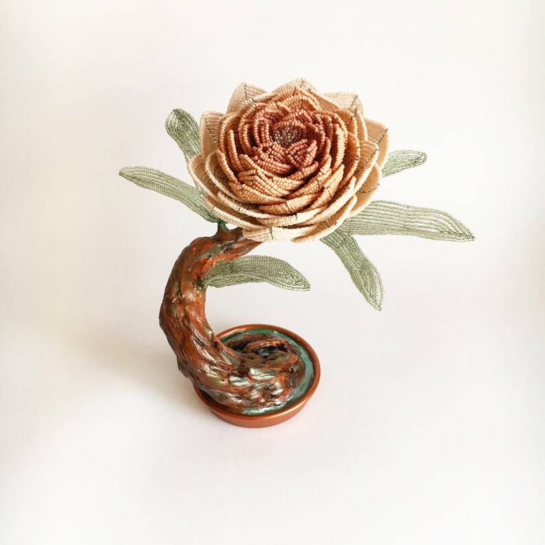 May include: A handcrafted, beaded flower sculpture with a detailed, layered petal design in shades of beige and brown. The flower sits atop a curved, textured, brown stem and a copper-colored base with a touch of green.