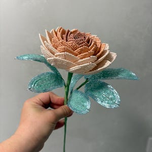 May include: A handcrafted flower sculpture made from small beads. The petals transition from a deep peach at the center to a pale cream at the edges. The stem and leaves are constructed from vibrant turquoise beads, creating a contrast with the flower.