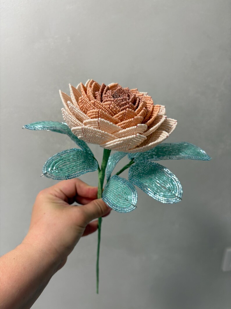 May include: A handcrafted flower sculpture made from small beads. The petals transition from a deep peach at the center to a pale cream at the edges. The stem and leaves are constructed from vibrant turquoise beads, creating a contrast with the flower.