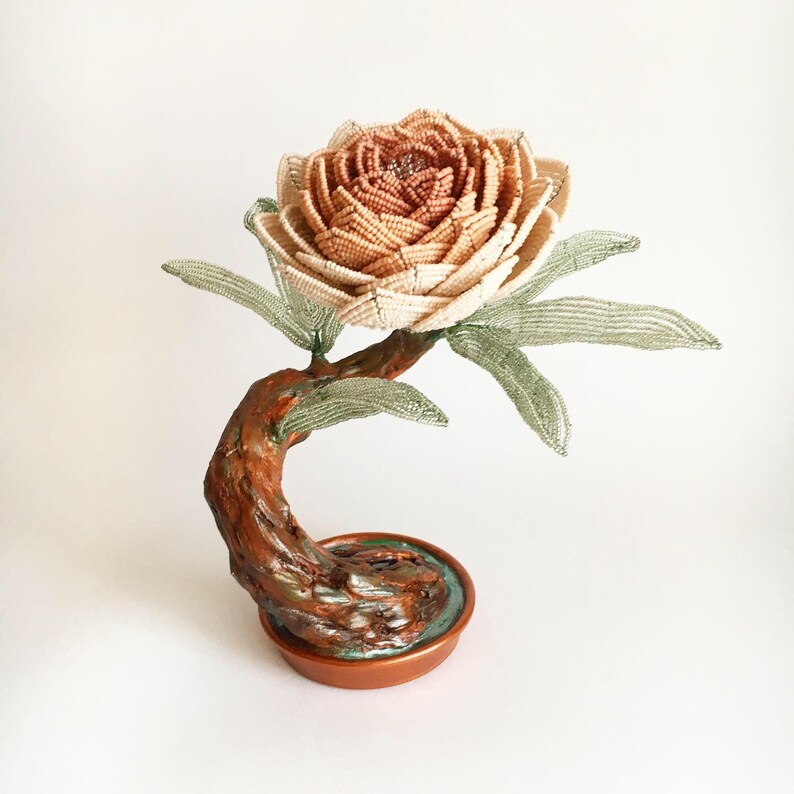 May include: A handcrafted beaded flower sculpture in shades of beige and peach, with green beaded leaves. The flower sits atop a brown, curved stem and a copper-colored base, resembling a bonsai tree.