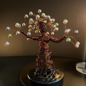 May include: A handmade wire sculpture of a figure with outstretched arms, decorated with pearl-like beads. The sculpture is made of copper-coloured wire and sits on a black base. The beads are in shades of white, pink, and green.