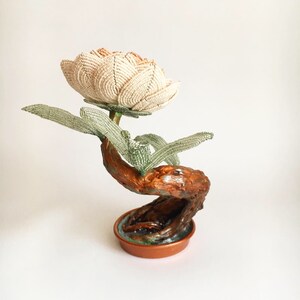 May include: A handcrafted beaded flower sculpture. The flower is cream-colored with a touch of peach, and the leaves are green. The stem and base are made of wood, painted in shades of brown and copper. The sculpture is a unique decorative item.