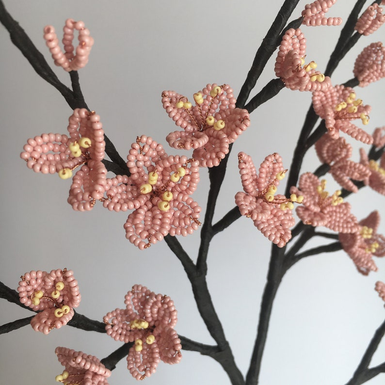 May include: A delicate beaded floral arrangement featuring pink and yellow blossoms on black wire branches. The flowers are crafted from small beads, creating a realistic and intricate design.