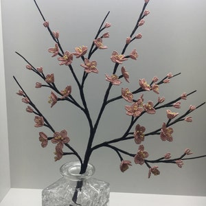 May include: A delicate pink beaded flower arrangement in a clear glass vase. The flowers are arranged in a branch-like shape, creating a realistic and elegant display.
